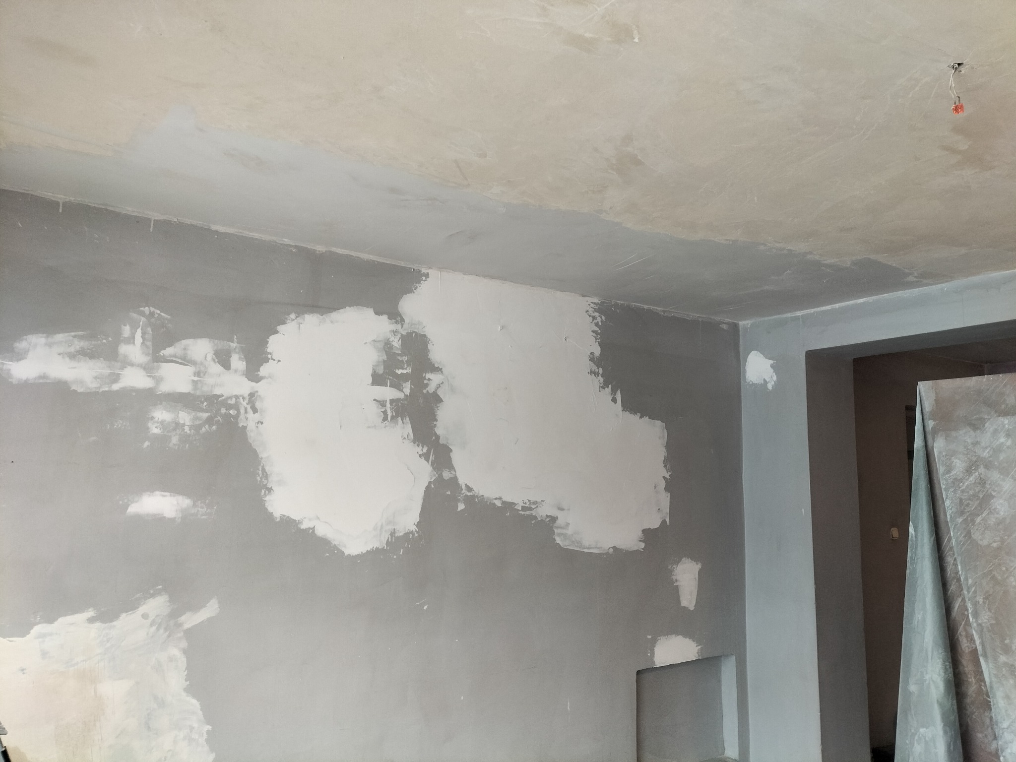 Ściana w trakcie przygotowania do malowania, z widocznymi łatami świeżo nałożonej masy szpachlowej, sufit częściowo pomalowany na jasny kolor, widoczne przewody elektryczne.