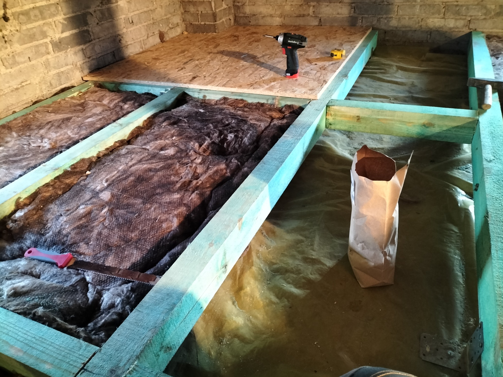 Konstrukcja podłogi z drewnianych belek, wypełniona brązową wełną mineralną, częściowo przykryta płytą OSB, w tle wiertarko-wkrętarka Metabo i miarka, obok papierowy worek z materiałem izolacyjnym...