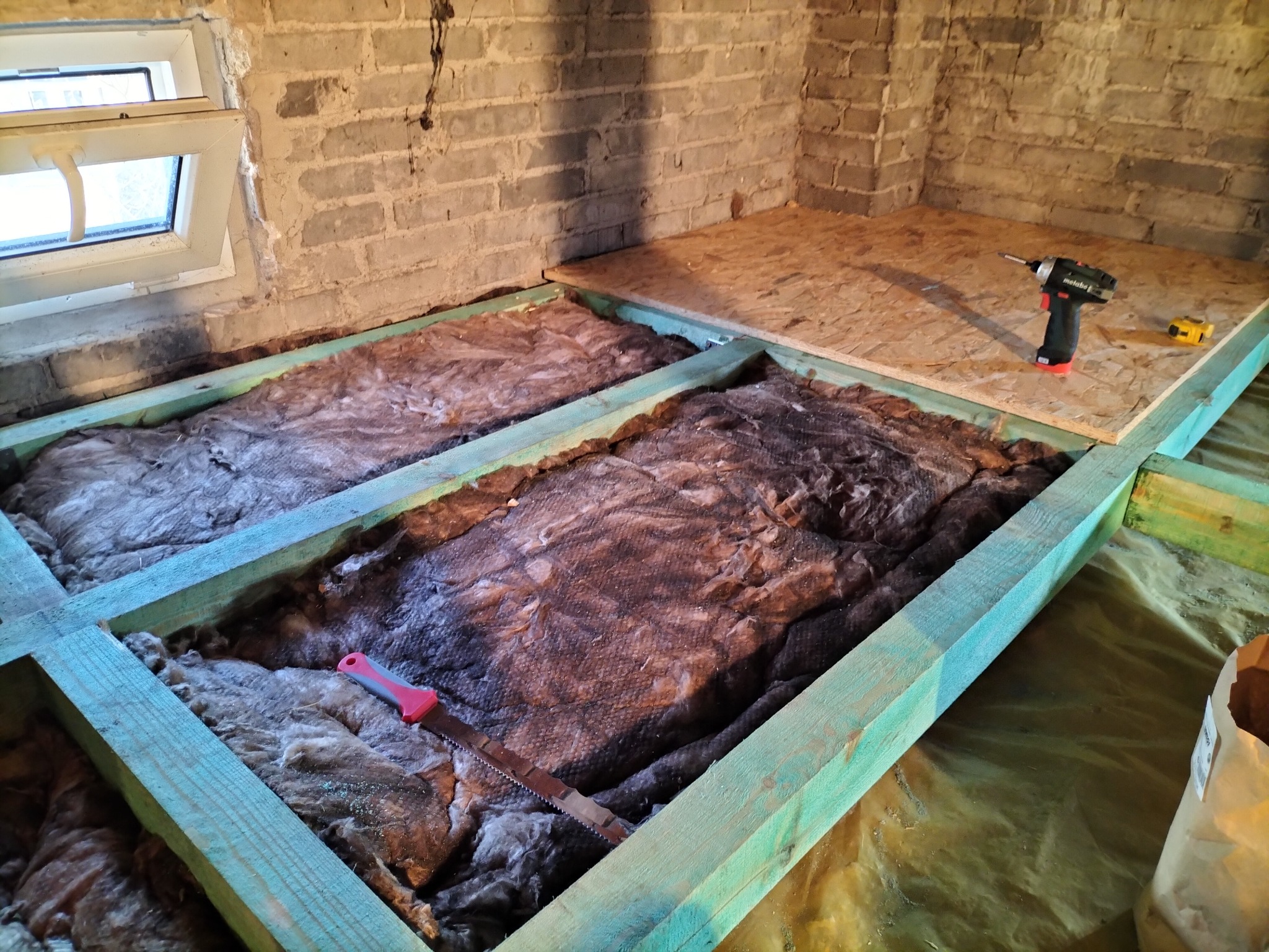 Konstrukcja drewniana z izolacją z wełny mineralnej, częściowo przykryta płytą OSB, w tle ceglana ściana z oknem, wiertarka akumulatorowa i miara zwijana na płycie, piła ręczna z czerwonym uchwytem...