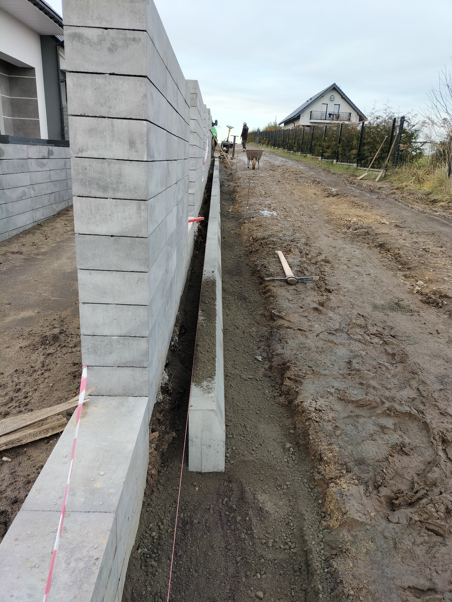 Budowa ogrodzenia z prefabrykatów betonowych: widok na fragment muru z szarego betonu, wykop z widoczną linką wyznaczającą linię, w tle pracownicy i domy.