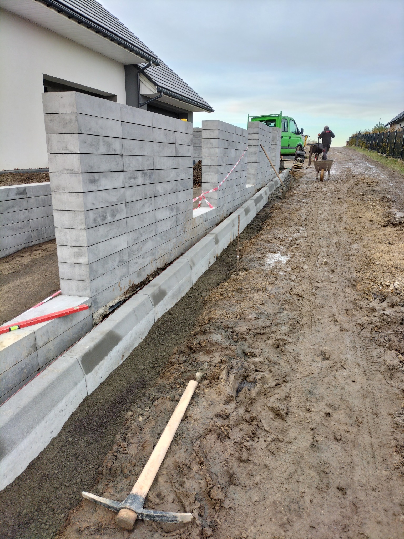 Budowa ogrodzenia z betonowych bloczków i krawężników; widoczny poziomica, wykop, ziemia i pracownik z taczką w tle.