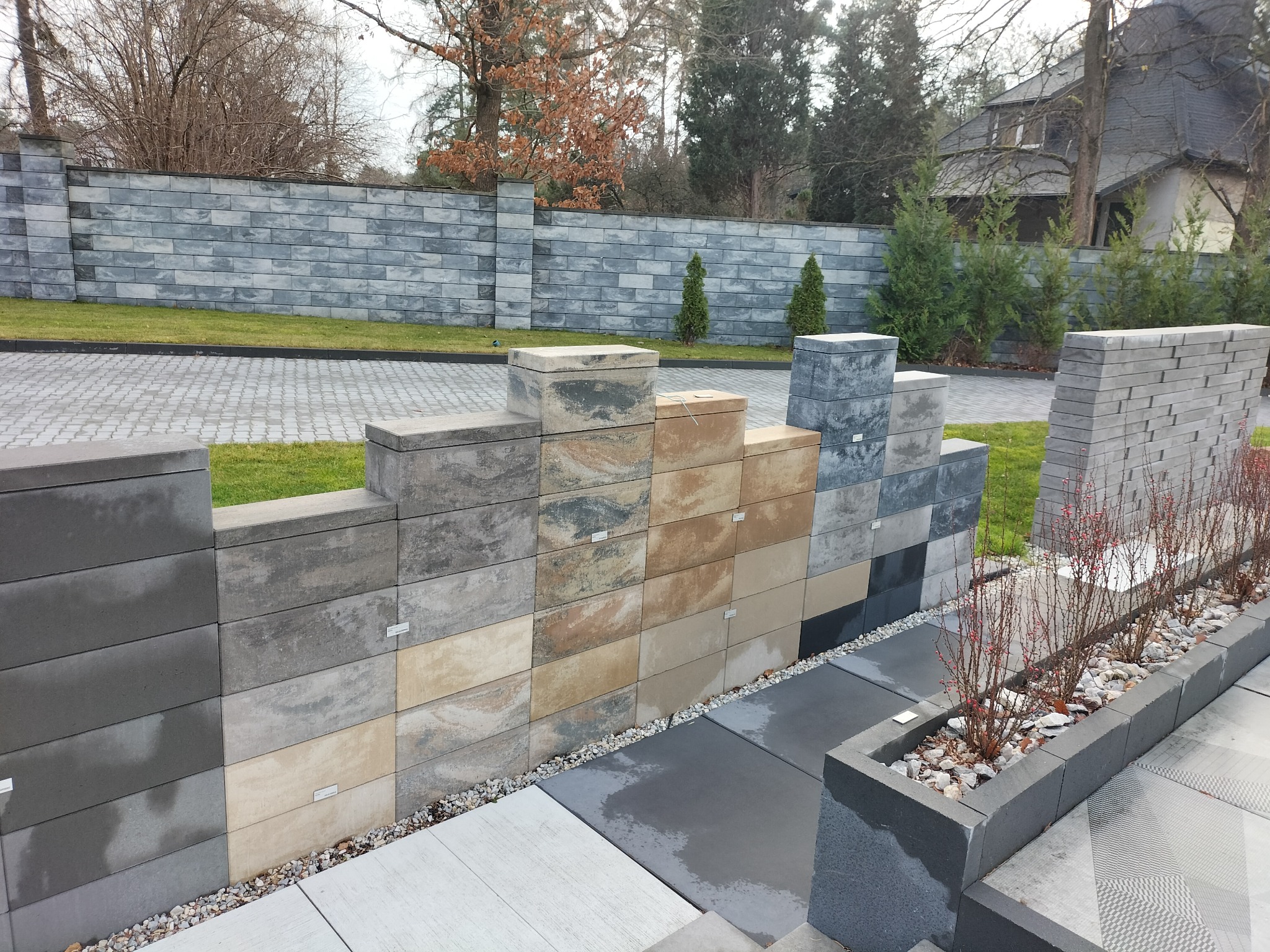 Ekspozycja różnych kolorów i faktur bloczków betonowych na tle ogrodzenia z tego samego materiału, prezentacja aranżacji przestrzeni zewnętrznej.