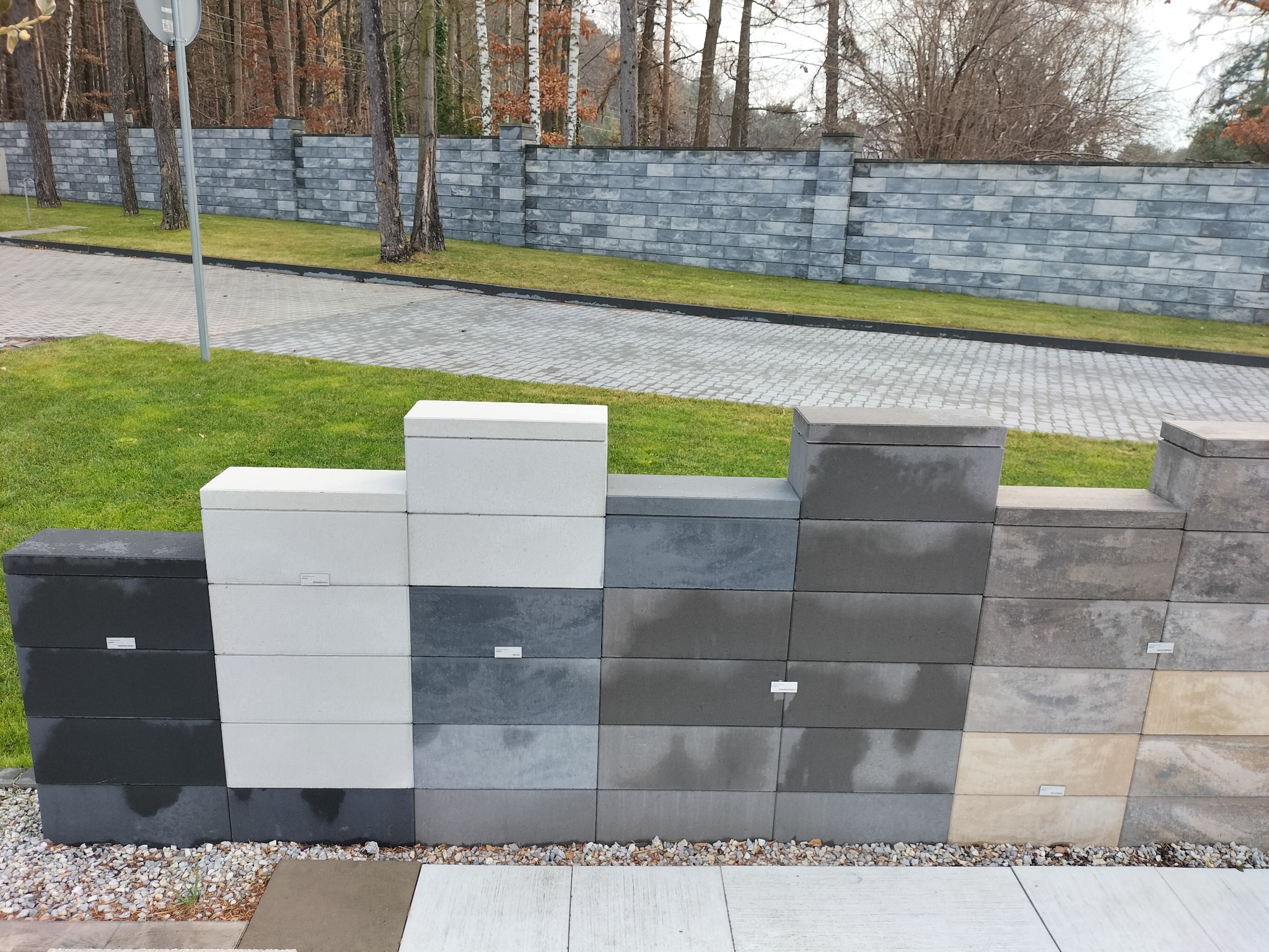 Ekspozycja różnokolorowych bloczków betonowych na zewnątrz, na tle zielonego trawnika i szarego muru z podobnych elementów.