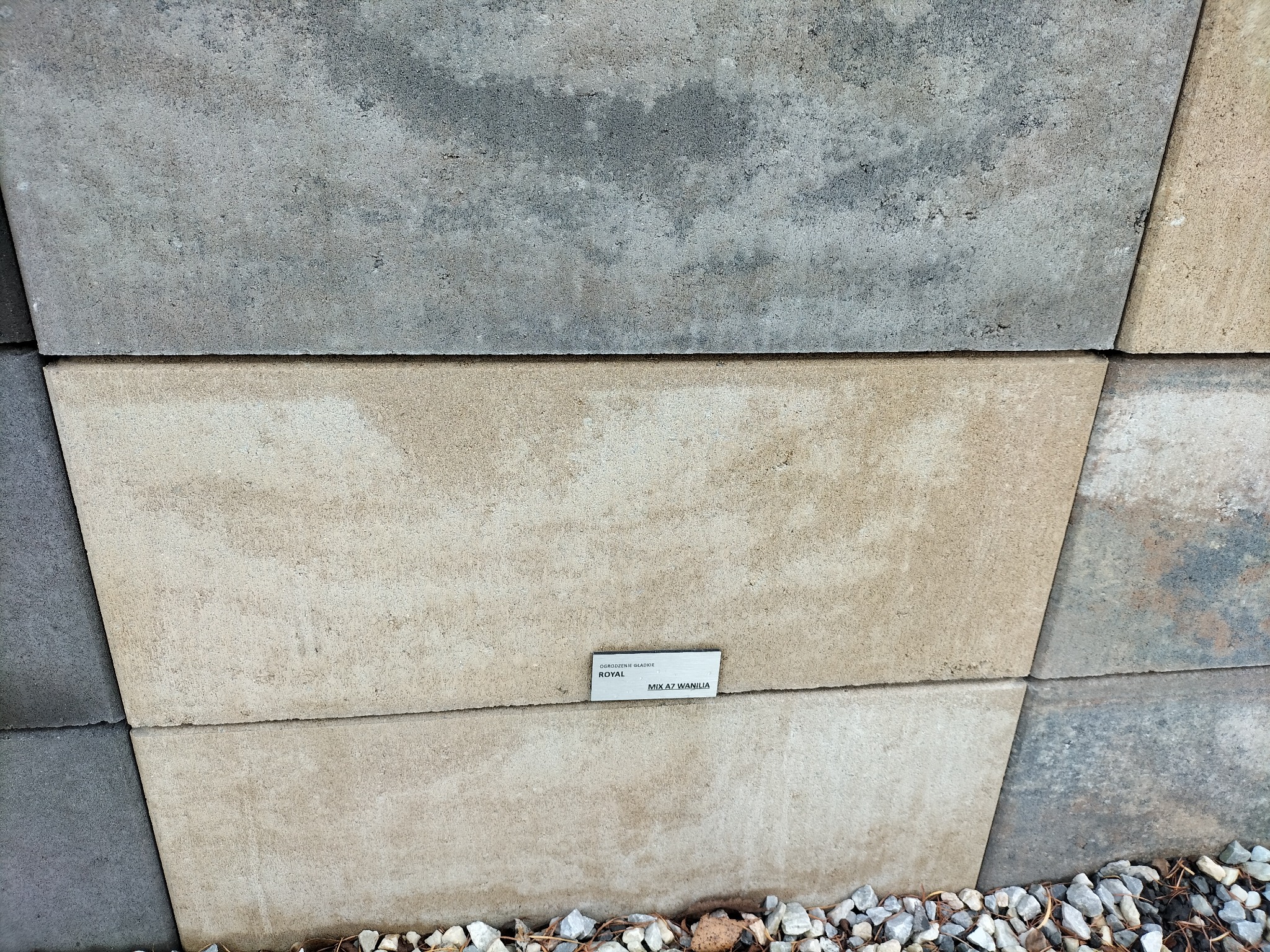 Prezentacja kostki brukowej w odcieniach szarości i beżu z drobnymi kamieniami u podstawy i etykietą informacyjną na jednej z kostek.