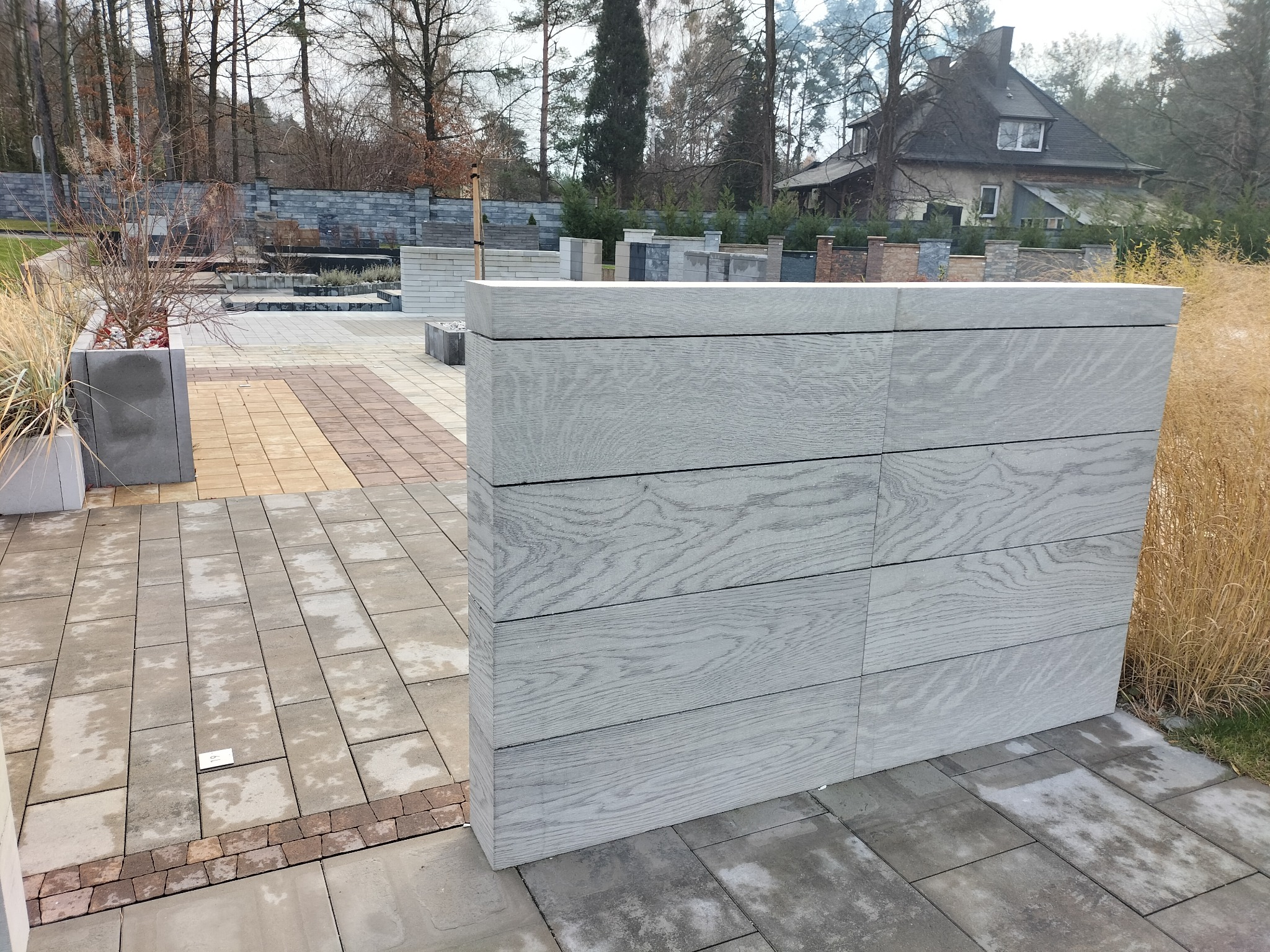 Nowoczesny szary murek z paneli imitujących drewno na tle posesji z kostką brukową i roślinnością, w tle widoczny dom.