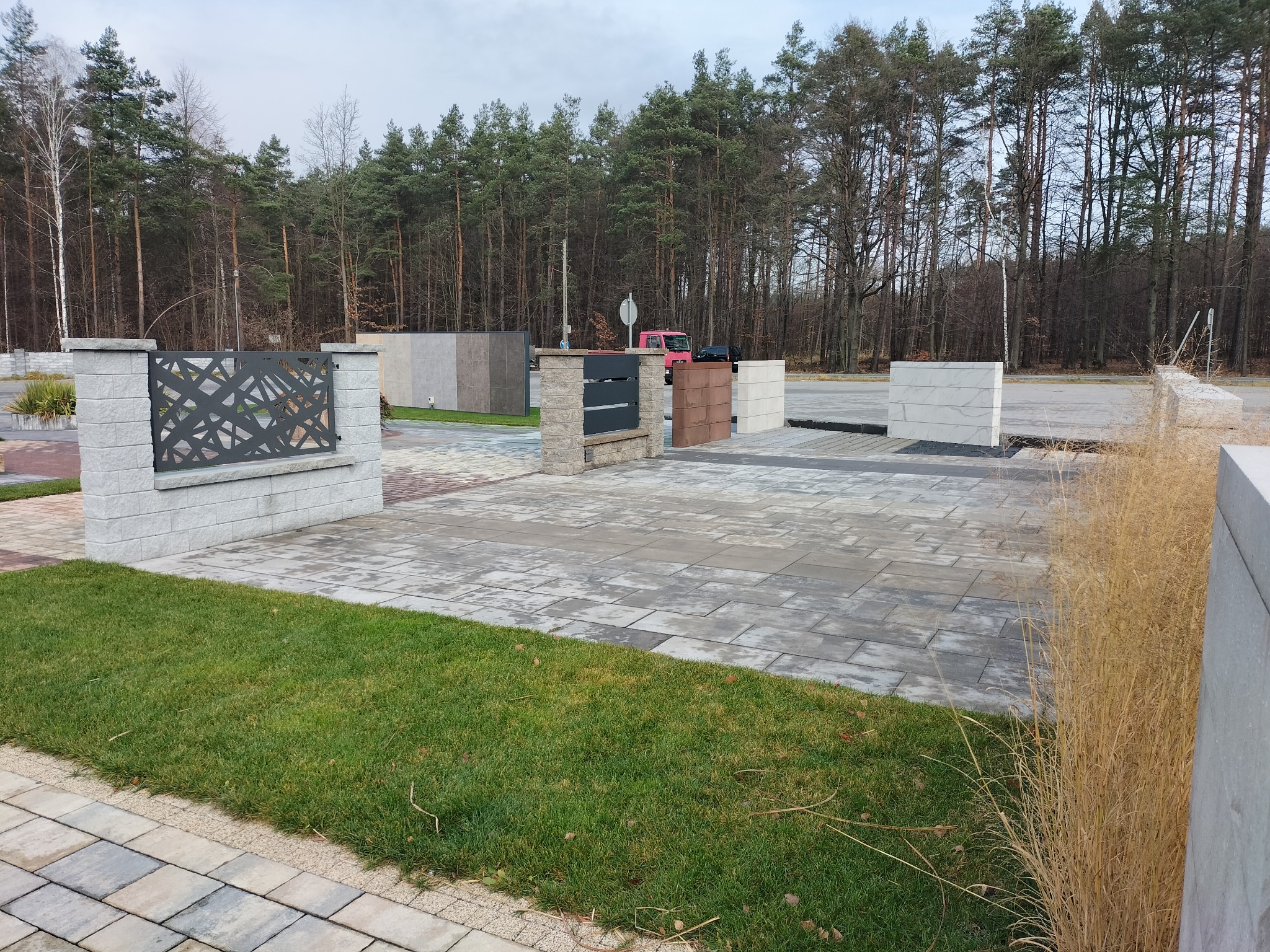 Wystawa kostki brukowej i elementów ogrodzeniowych z betonu architektonicznego, prezentująca różne wzory, kolory i faktury na tle lasu.