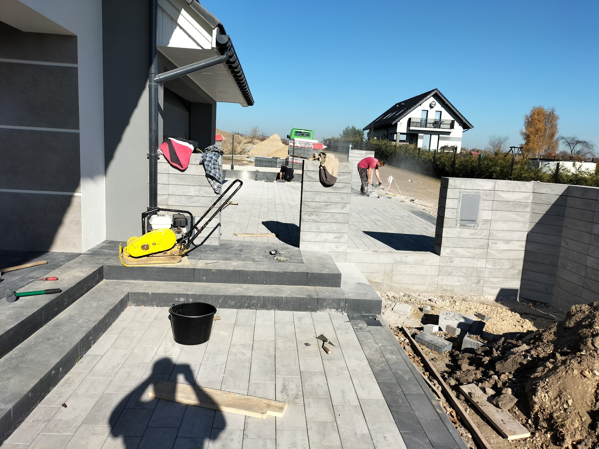 Prace brukarskie w trakcie: układanie kostki brukowej na posesji przed domem jednorodzinnym, widoczny pracownik docinający elementy, zagęszczarka, narzędzia i fragment ułożonego chodnika z kostki.