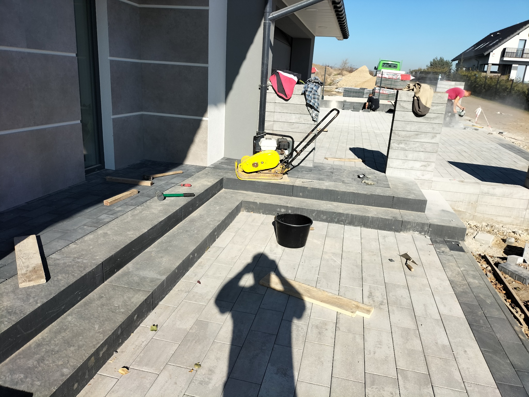 Montaż kostki brukowej na schodach wejściowych do domu, widoczna ubijarka wibracyjna, czarne wiadro oraz cień osoby robiącej zdjęcie, w tle pracownicy układający kostkę.