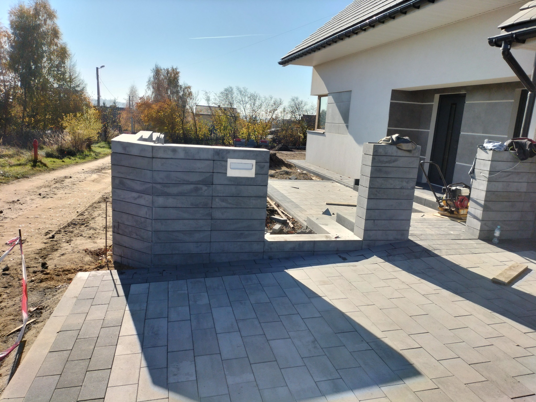 Ułożona szara kostka brukowa na podjeździe przed domem z widocznym ogrodzeniem z szarej kostki, w tle drzewa z jesiennymi liśćmi.