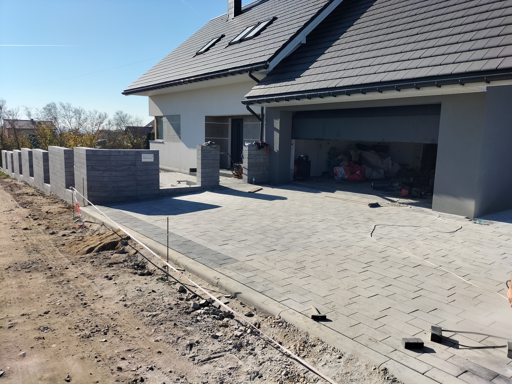 Nowoczesny dom jednorodzinny z szarą kostką brukową na podjeździe i chodniku, ogrodzenie z betonowych bloczków, widoczny teren po robotach ziemnych.