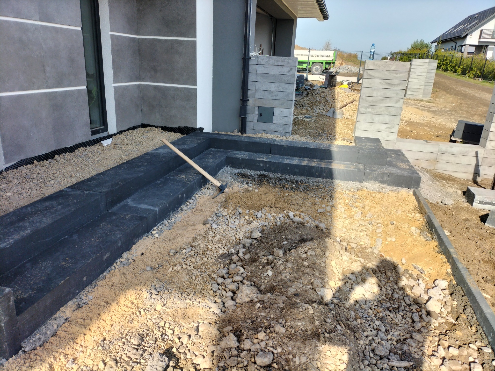 Budowa schodów zewnętrznych z betonowych elementów, widoczne przygotowane podłoże z piasku i kamieni, w tle budynek mieszkalny i ciężarówka.