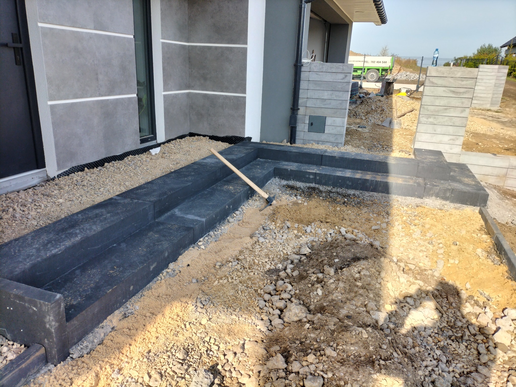 Budowa schodów wejściowych z ciemnych betonowych bloczków, widoczne narzędzie - łopata, teren wokół wysypany żwirem i piaskiem, fragment elewacji budynku z szarymi panelami.