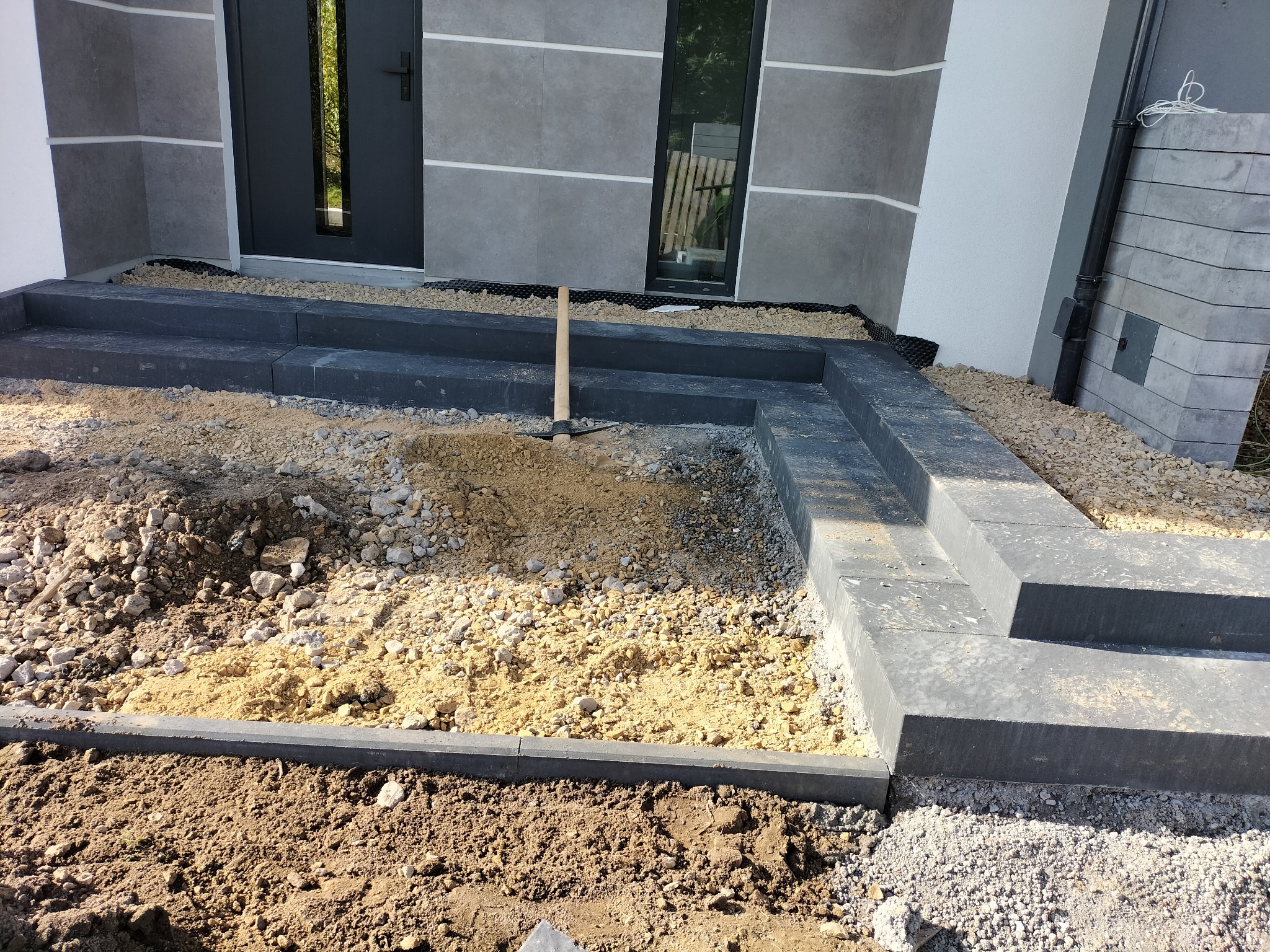 Montaż schodów zewnętrznych z ciemnoszarych prefabrykatów betonowych; widoczny etap przygotowania podłoża z piasku i gruzu, w tle ściana budynku z szarych płyt i czarne drzwi wejściowe, leżące obok...