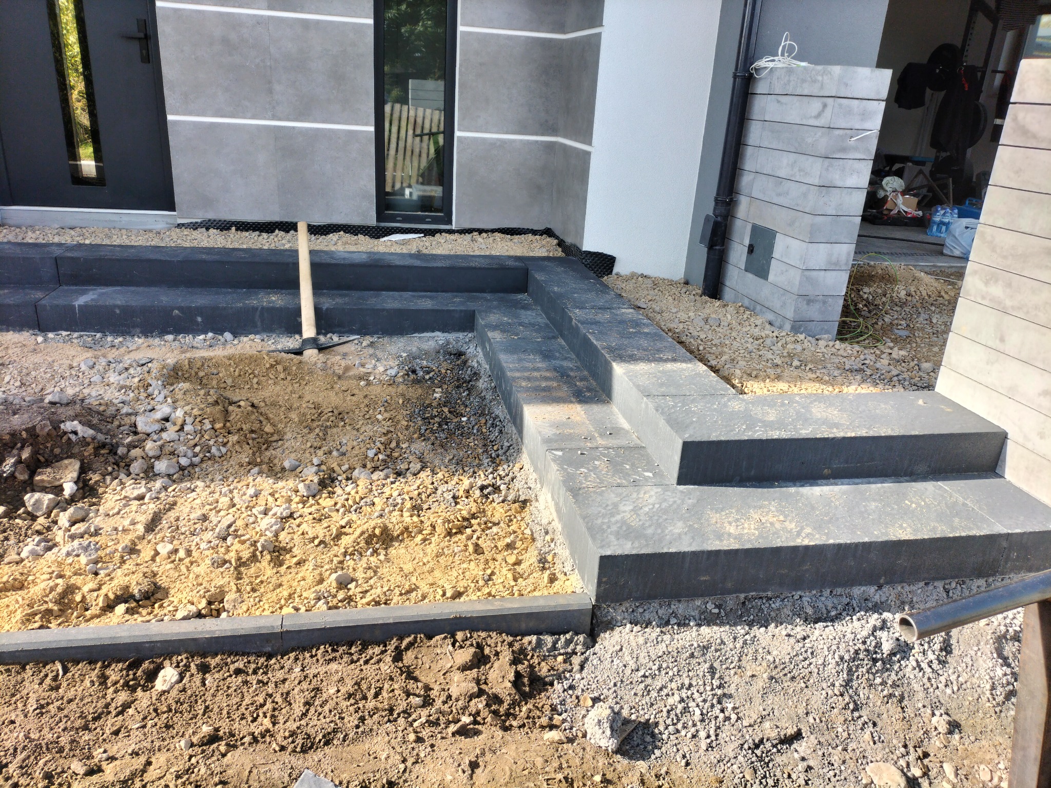 Montaż schodów zewnętrznych z betonowych bloków w Łazach Śląskich, widoczny etap przygotowania podłoża z użyciem tłucznia i piasku, obok budynku z szarą elewacją i stolarką okienną.