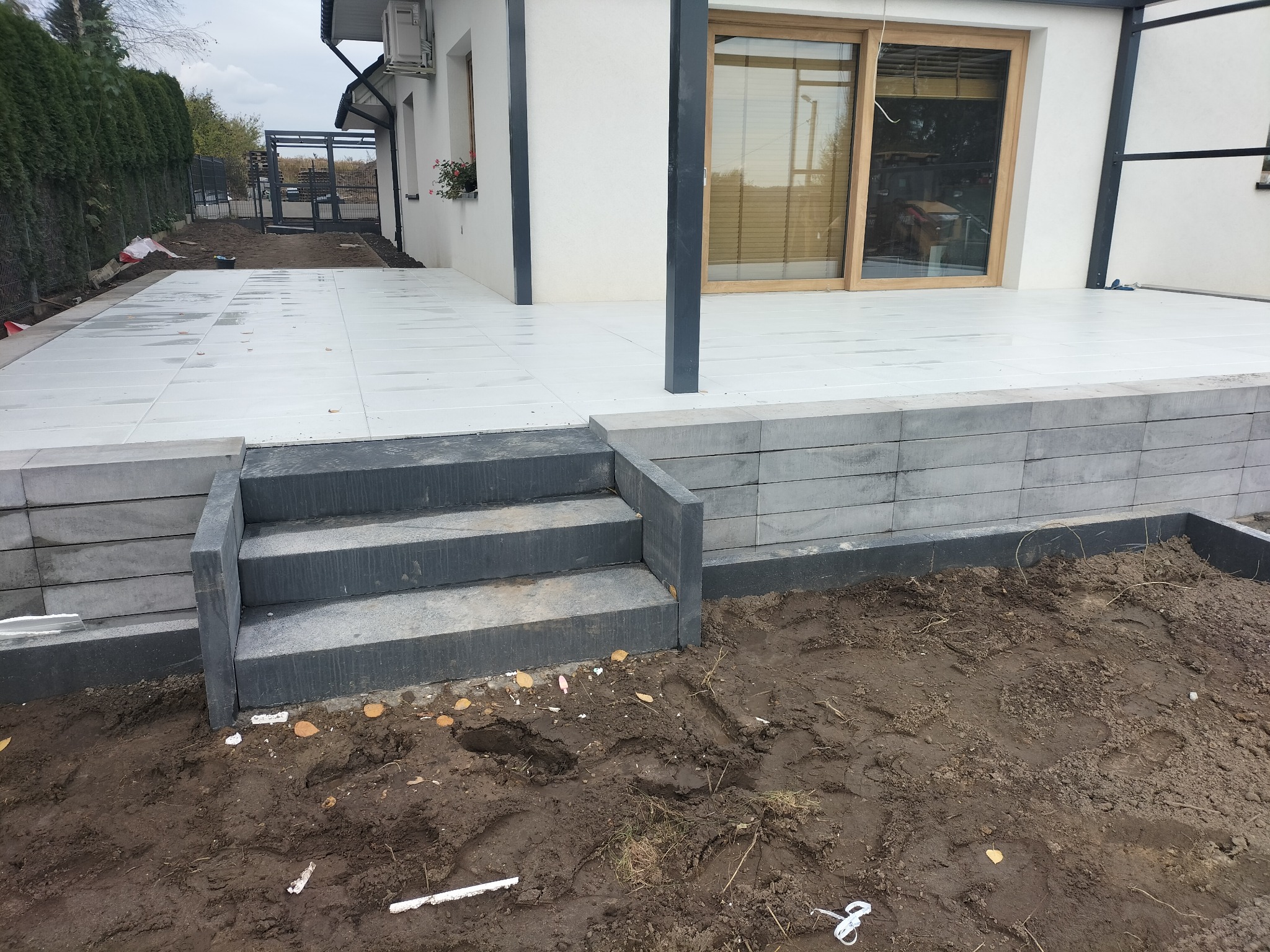 Nowoczesny taras z jasnych płyt, z trzema schodami z szarej kostki, widok na ogród i fragment domu z oknem przesuwnym.