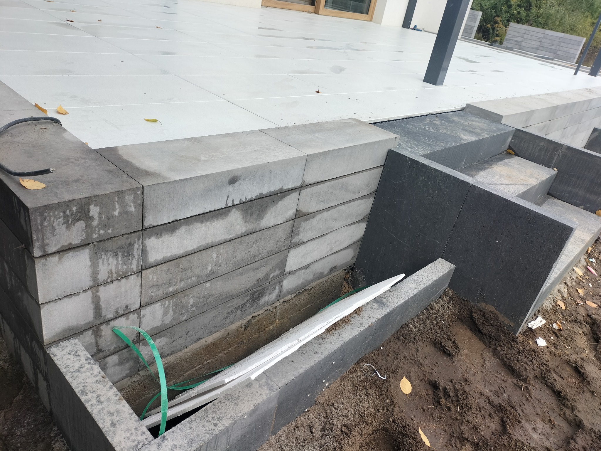 Montaż obrzeży betonowych przy tarasie z płyt, widoczne bloczki betonowe, zielona taśma miernicza i fragmenty białego styropianu w wykopie.