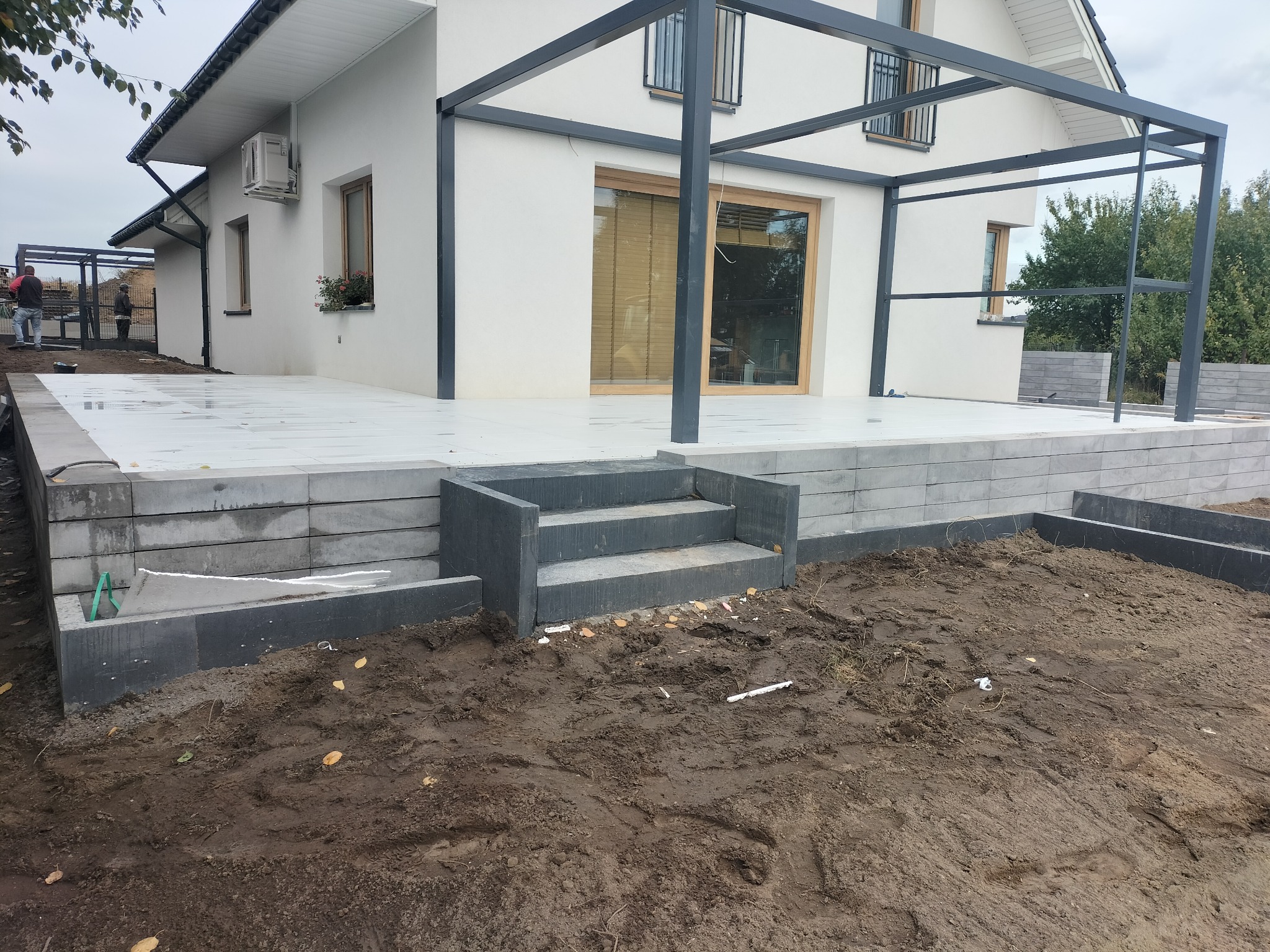 Nowoczesny taras z jasnoszarych płyt, schody z kostki brukowej i metalowa pergola przy białym domu jednorodzinnym, widoczne przygotowania do prac ziemnych w ogrodzie.