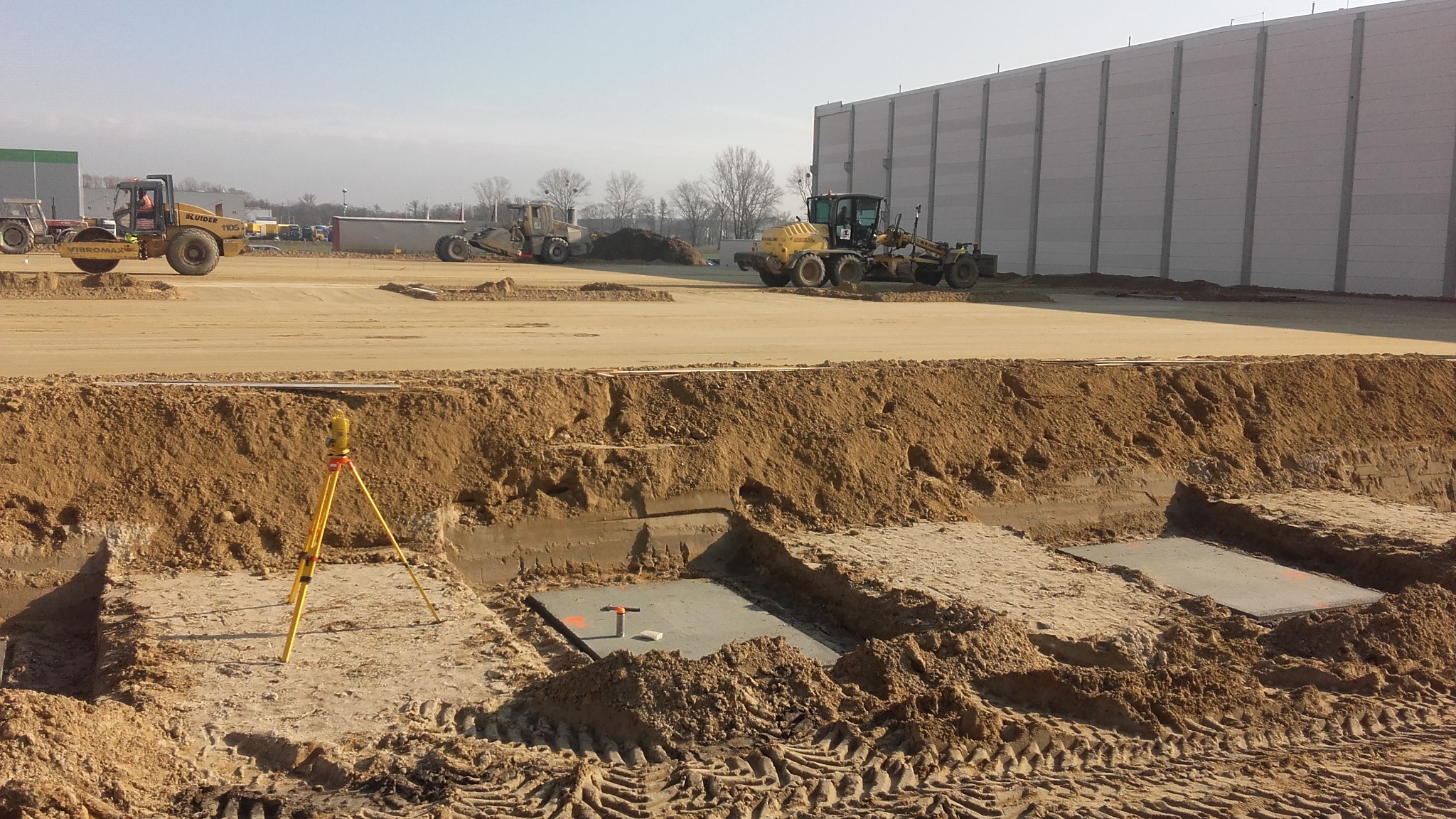 Przygotowany wykop pod fundament z betonową płytą, żółty niwelator optyczny na statywie, ciężki sprzęt budowlany w tle i hala magazynowa.