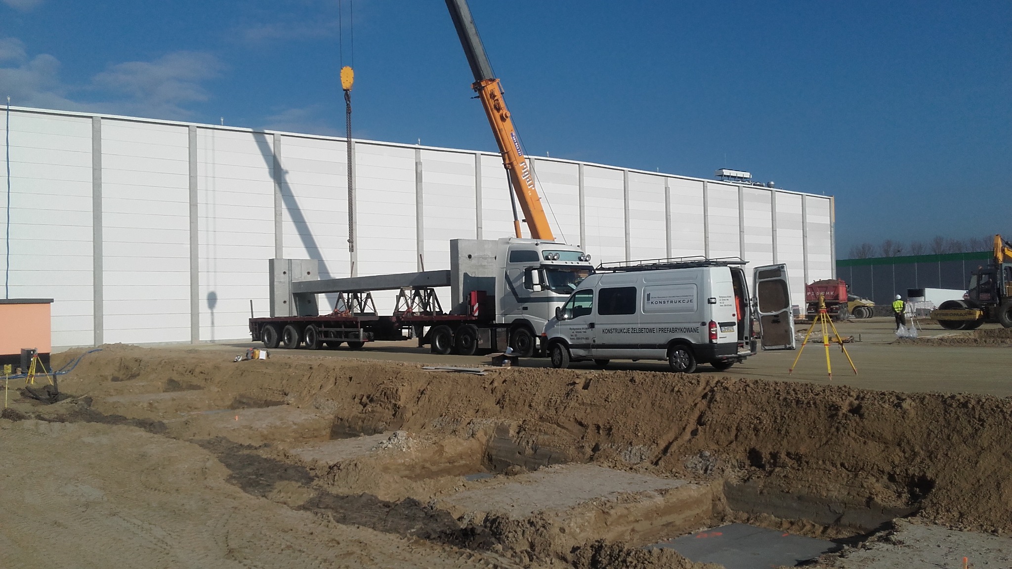 Montaż prefabrykowanych elementów betonowych na placu budowy, widoczny dźwig, ciężarówka z naczepą, wykopane fundamenty i geodeta z niwelatorem.