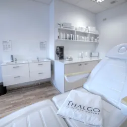 Biały fotel kosmetyczny z ręcznikiem marki Thalgo w jasnym, minimalistycznym gabinecie z umywalkami i p&oacute;łkami z kosmetykami.