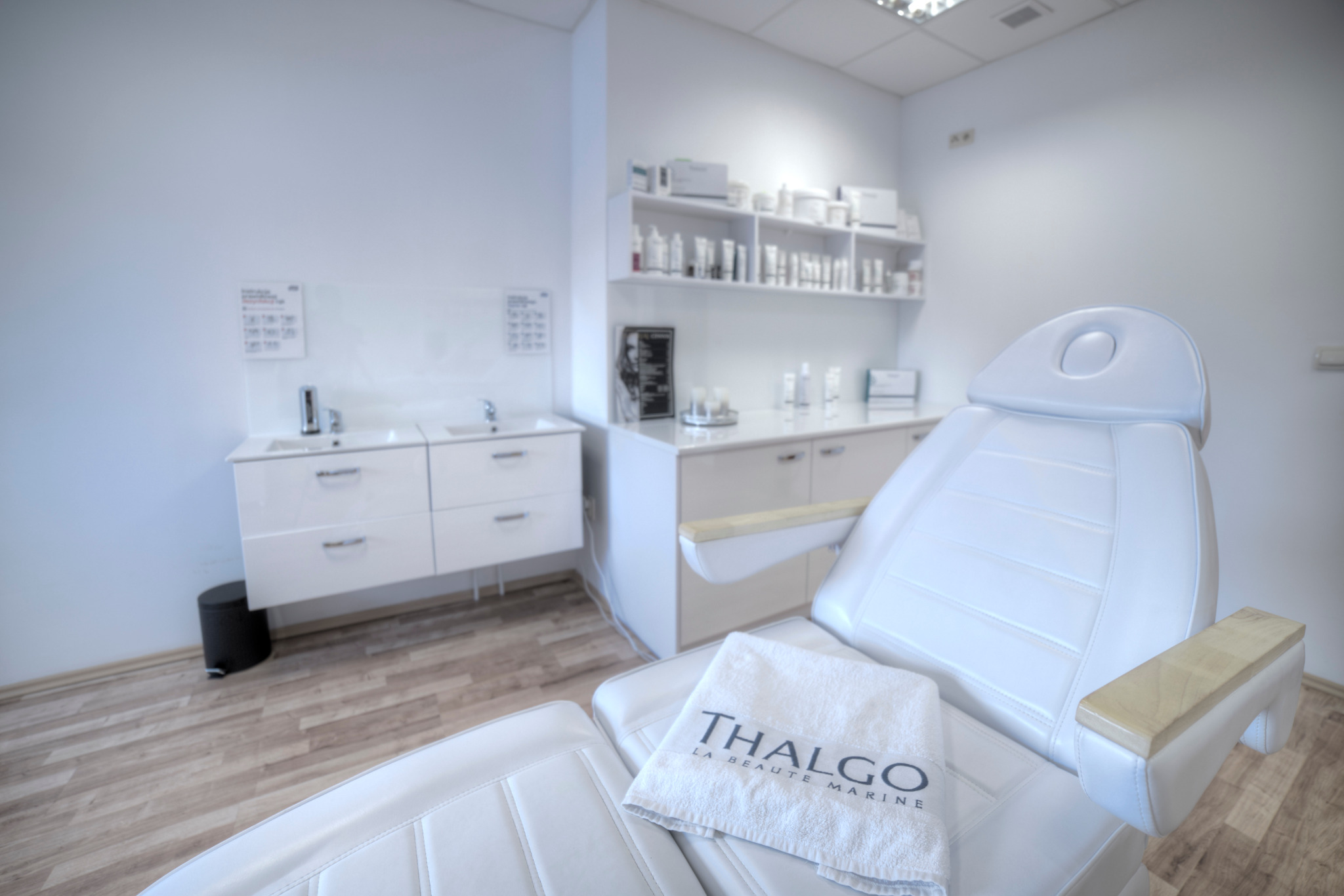 Biały fotel kosmetyczny z ręcznikiem marki Thalgo w jasnym, minimalistycznym gabinecie z umywalkami i półkami z kosmetykami.