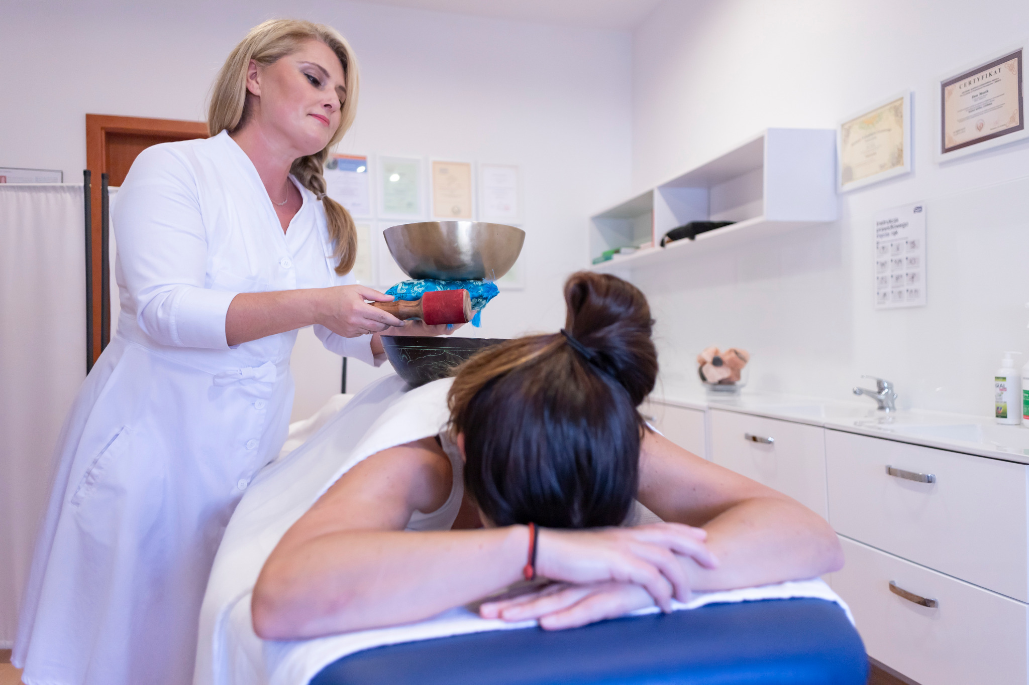 Klientka leżąca na stole do masażu, terapeutka w białym uniformie przygotowuje misy tybetańskie do terapii dźwiękiem, w tle widoczne certyfikaty na ścianie i białe szafki.