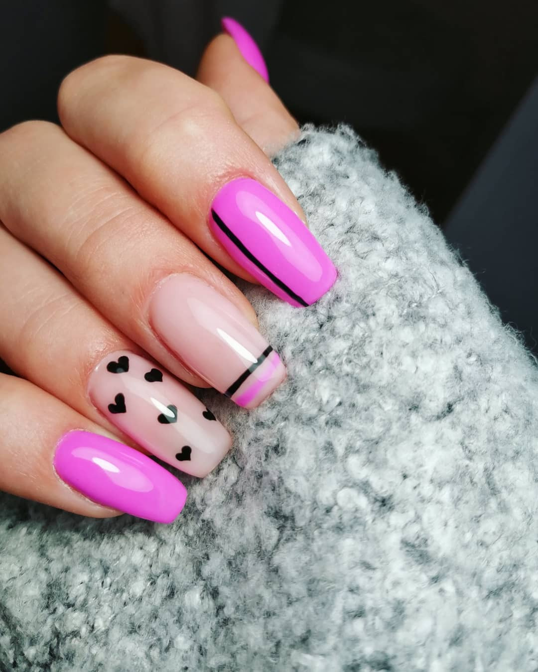 Dłoń z pomalowanymi paznokciami: trzy w kolorze fuksji, jeden z czarnymi sercami, jeden z czarnym paskiem, na tle szarego swetra.