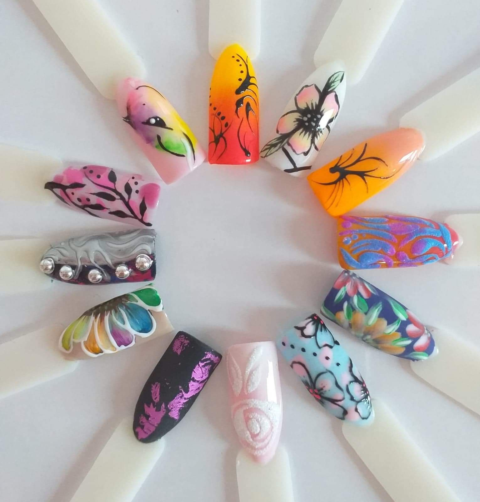 Prezentacja różnorodnych wzorów zdobień paznokci na plastikowych wzornikach ułożonych w okrąg, ukazująca kwiatowe motywy, abstrakcyjne wzory i efekty specjalne, takie jak folia i cyrkonie.
