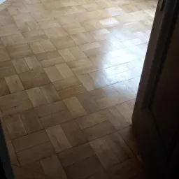 Parkiet ułożony w jodełkę w pustym pokoju z grzejnikiem, widok zza częściowo otwartych drewnianych drzwi.