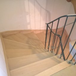 STYLDOM Mariusz Łukasiak - Jasne, drewniane schody z czarną, metalową balustradą widziane z góry, podczas remontu, z widocznymi śladami pyłu na podłodze.