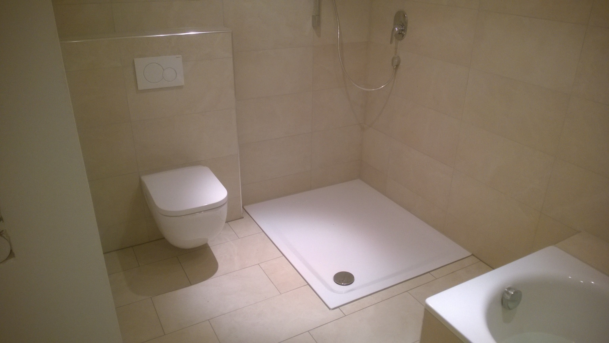 Jasna łazienka po remoncie: beżowe płytki, biała toaleta podtynkowa z przyciskiem dwudzielnym, brodzik i fragment wanny.