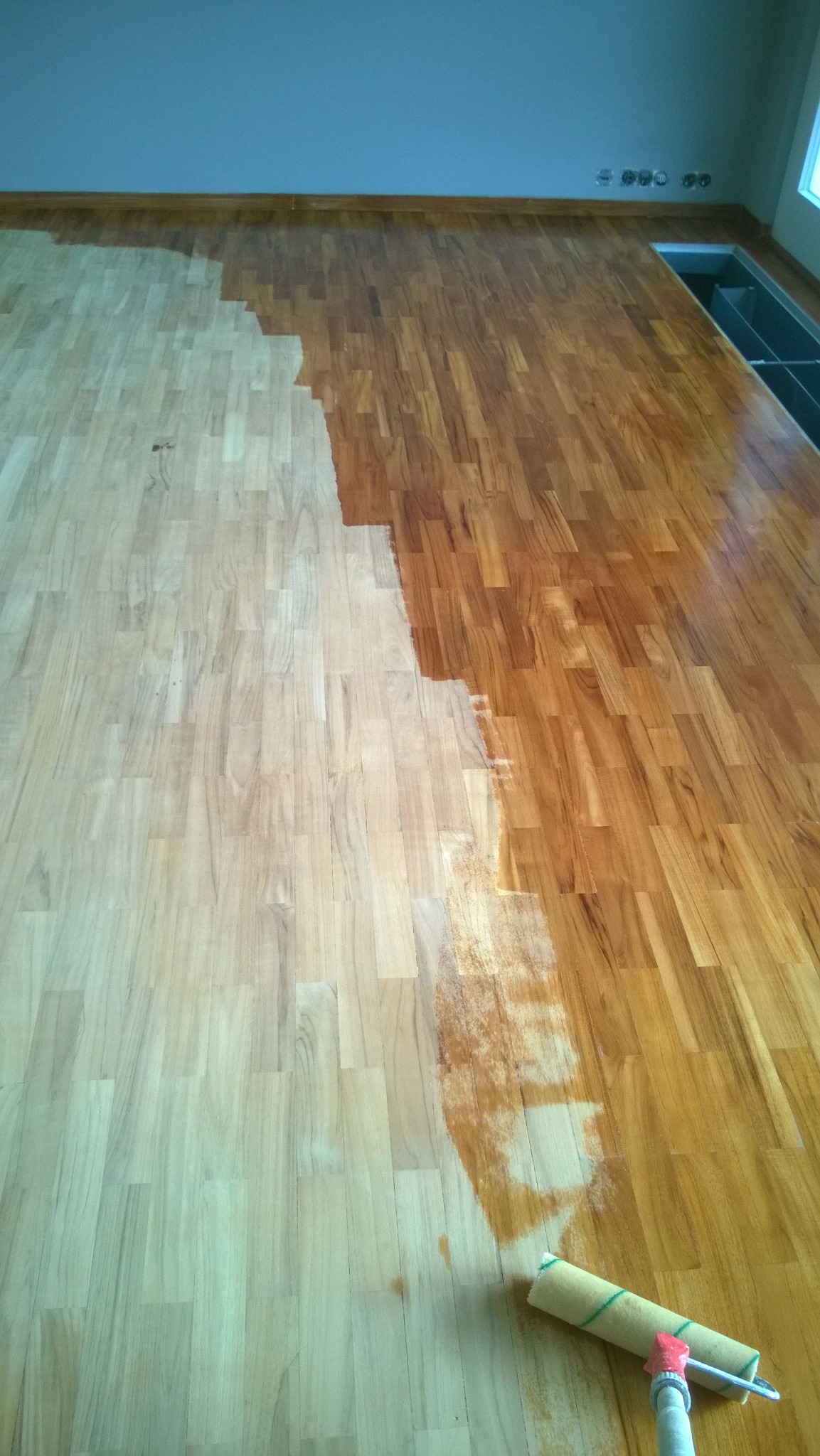 Częściowo polakierowany parkiet z widocznym wałkiem malarskim, ukazujący różnicę w kolorze przed i po lakierowaniu.