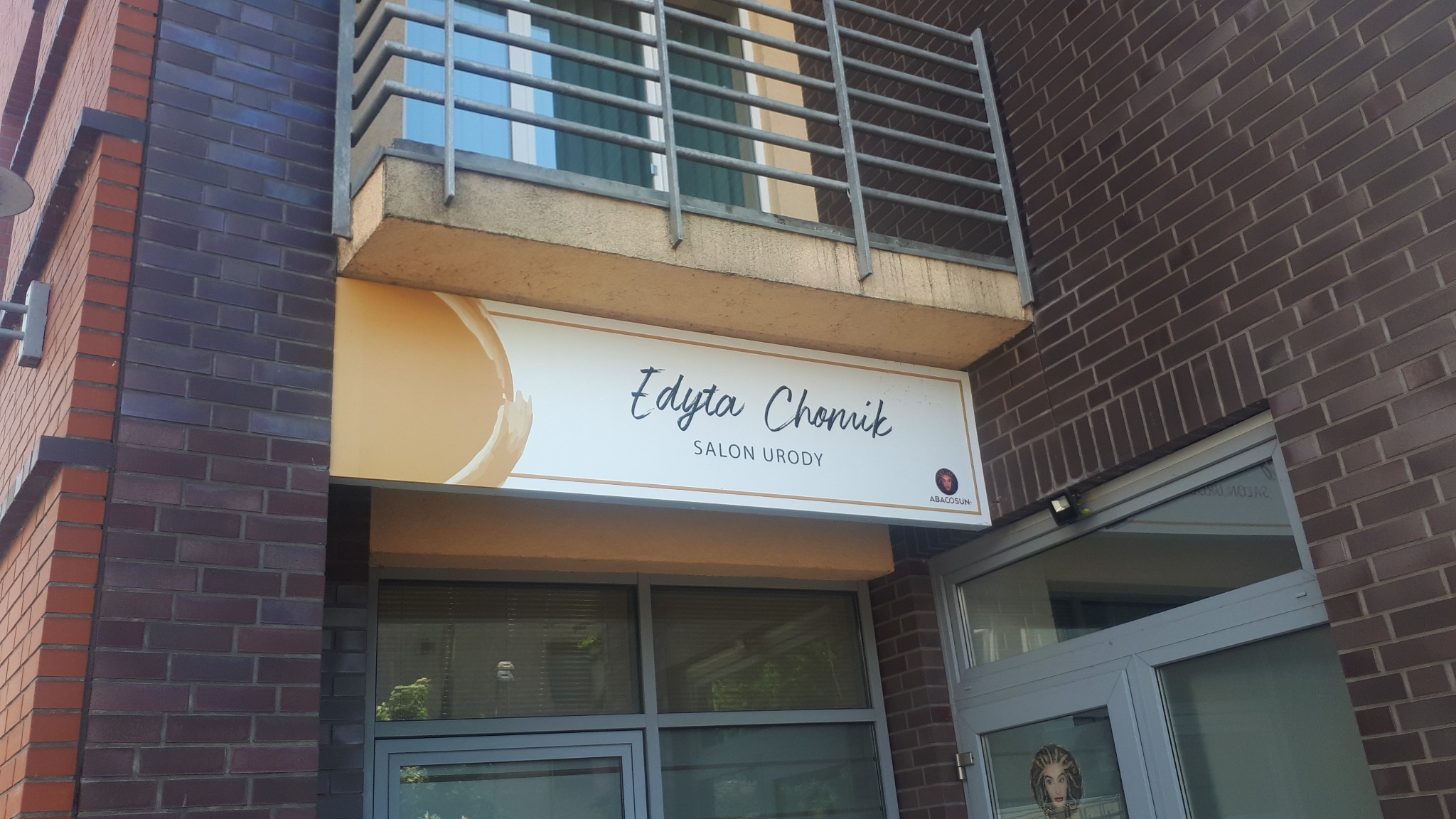 Szyld salonu urody 'Edyta Chonik' z logo Abacosun, zamontowany nad wejściem do budynku z cegły.