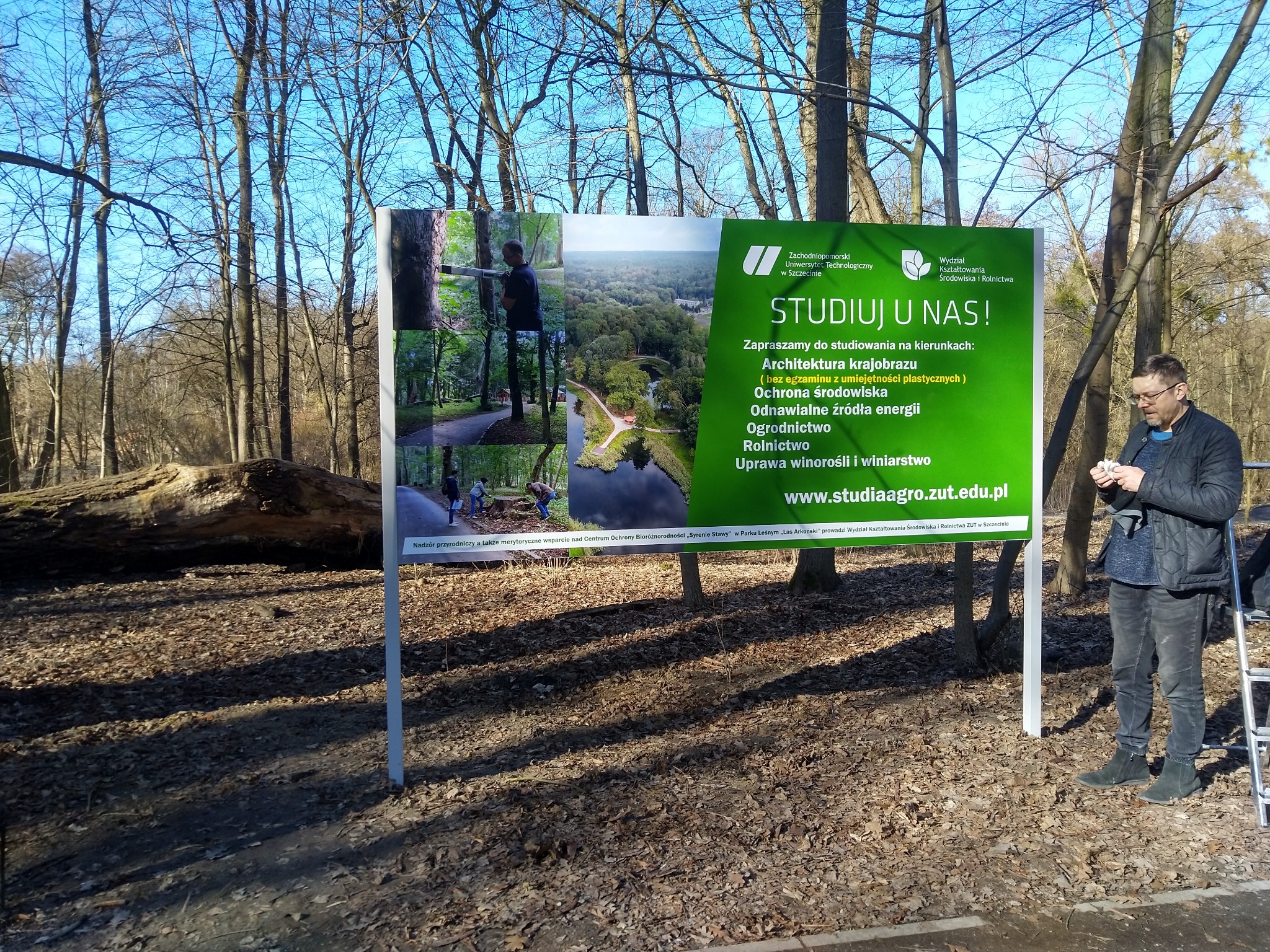 Zewnętrzny baner reklamowy uczelni z informacjami o kierunkach studiów, umieszczony w parku; mężczyzna w okularach przegląda coś na telefonie stojąc obok baneru.