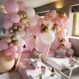 Dwa namioty tipi dla dzieci, udekorowane girlandami z różowych, białych i złotych balonów, z pluszowymi jednorożcami i poduszkami z cekinami w środku, ustawione na białych dywanach i stolikach.