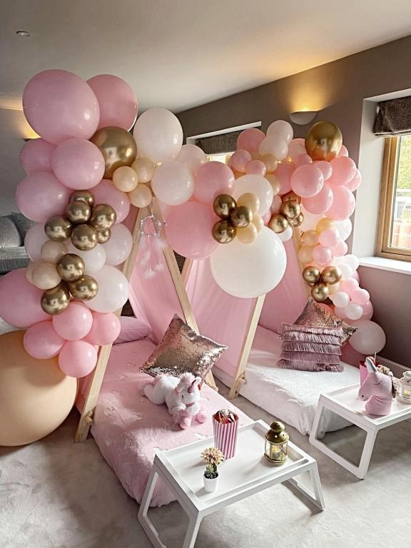 Dwa namioty tipi dla dzieci, udekorowane girlandami z różowych, białych i złotych balonów, z pluszowymi jednorożcami i poduszkami z cekinami w środku, ustawione na białych dywanach i stolikach.