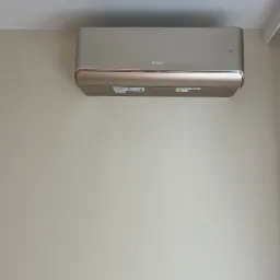 Zamontowany, beżowy klimatyzator marki Gree na jasnej ścianie. Widoczne detale urządzenia i fragment sufitu. Minimalistyczny design.