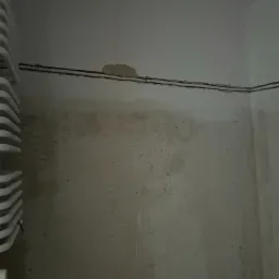 Biały, drabinkowy grzejnik łazienkowy zamontowany na ścianie z widocznymi śladami po instalacji elektrycznej w postaci przewodów w korytkach.