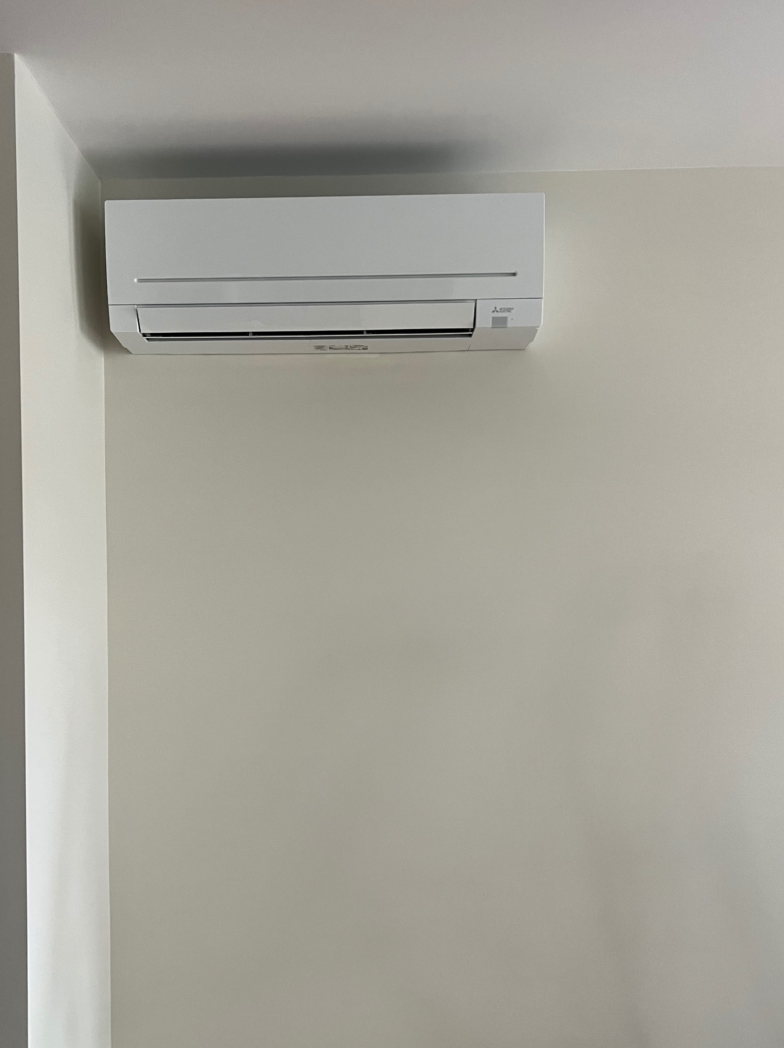 Biały klimatyzator Mitsubishi Electric zamontowany na jasnej ścianie w rogu pokoju. Minimalistyczny design, widoczne logo producenta.