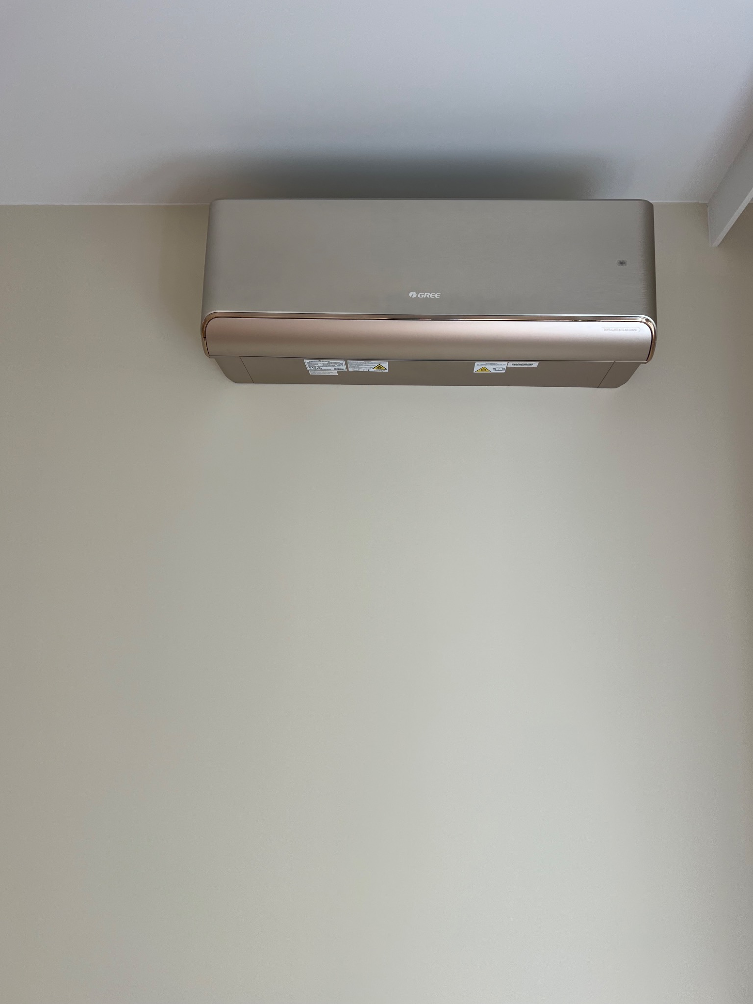 Zamontowany, beżowy klimatyzator marki Gree na jasnej ścianie. Widoczne detale urządzenia i fragment sufitu. Minimalistyczny design.