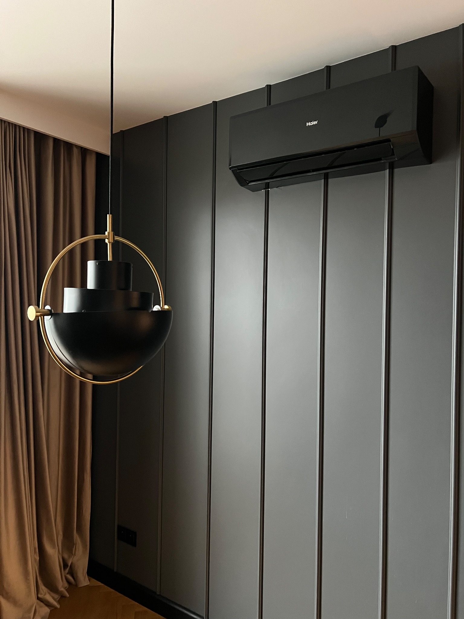 Czarny klimatyzator Haier na szarej, panelowej ścianie, obok designerska lampa wisząca z elementami złota i zasłona. Nowoczesne wnętrze.