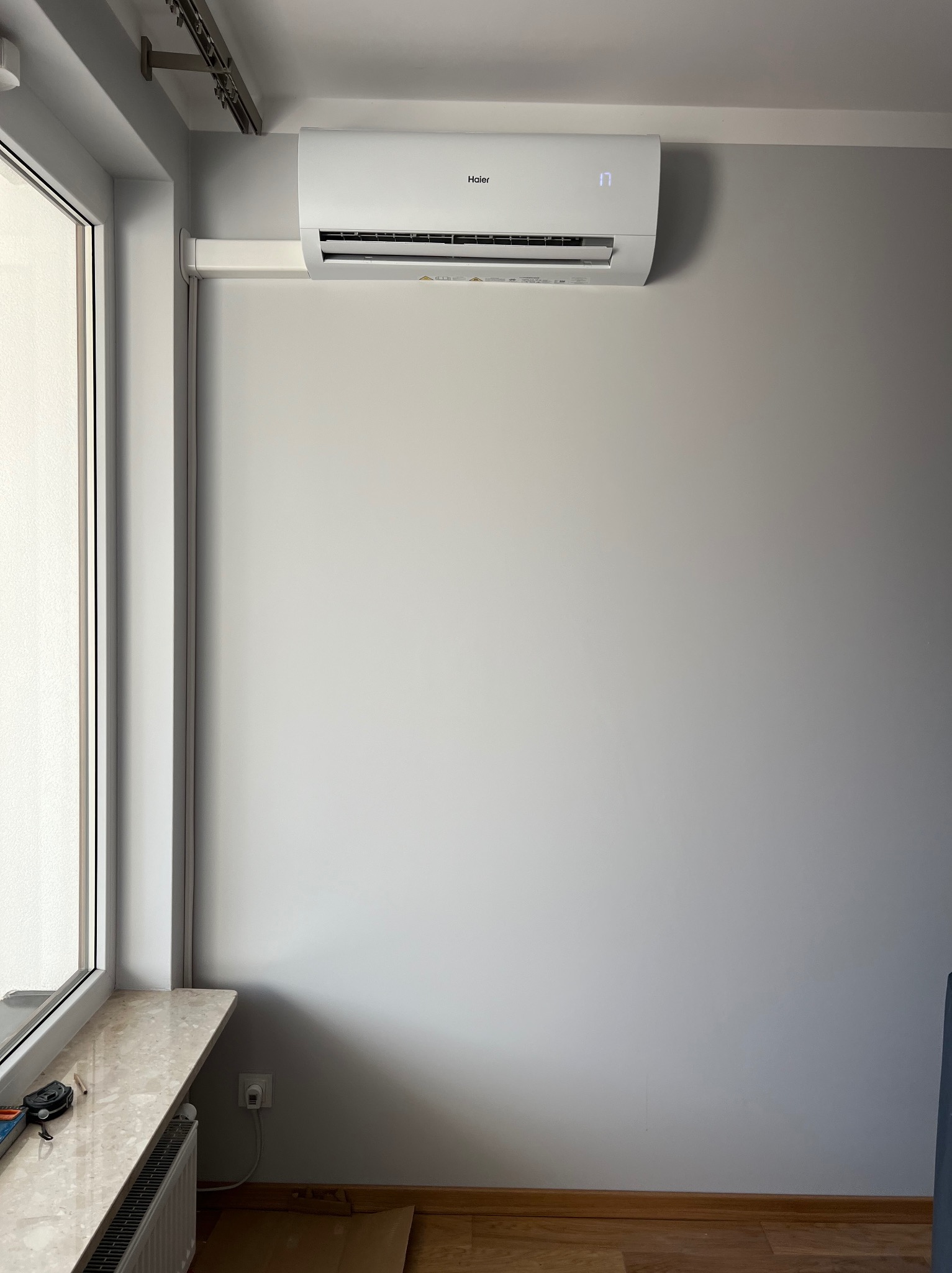 Biały klimatyzator Haier zamontowany na szarej ścianie w pomieszczeniu. Widoczny fragment okna z parapetem i grzejnikiem. Minimalistyczny design, nowoczesne wnętrze.