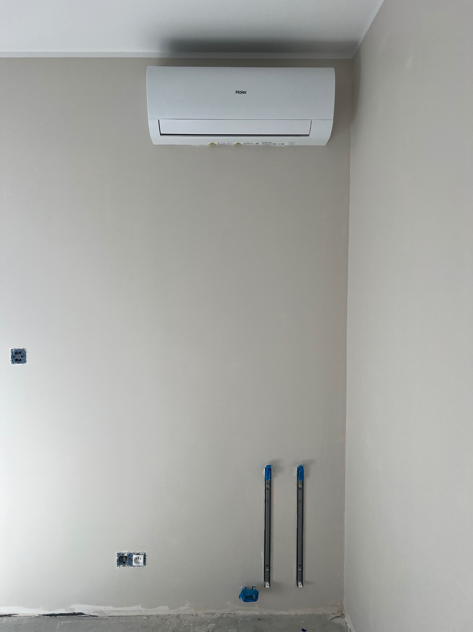 Biały klimatyzator Haier na ścianie w jasnym pokoju, widoczne przygotowane instalacje elektryczne i hydrauliczne. Świeżo pomalowane ściany.