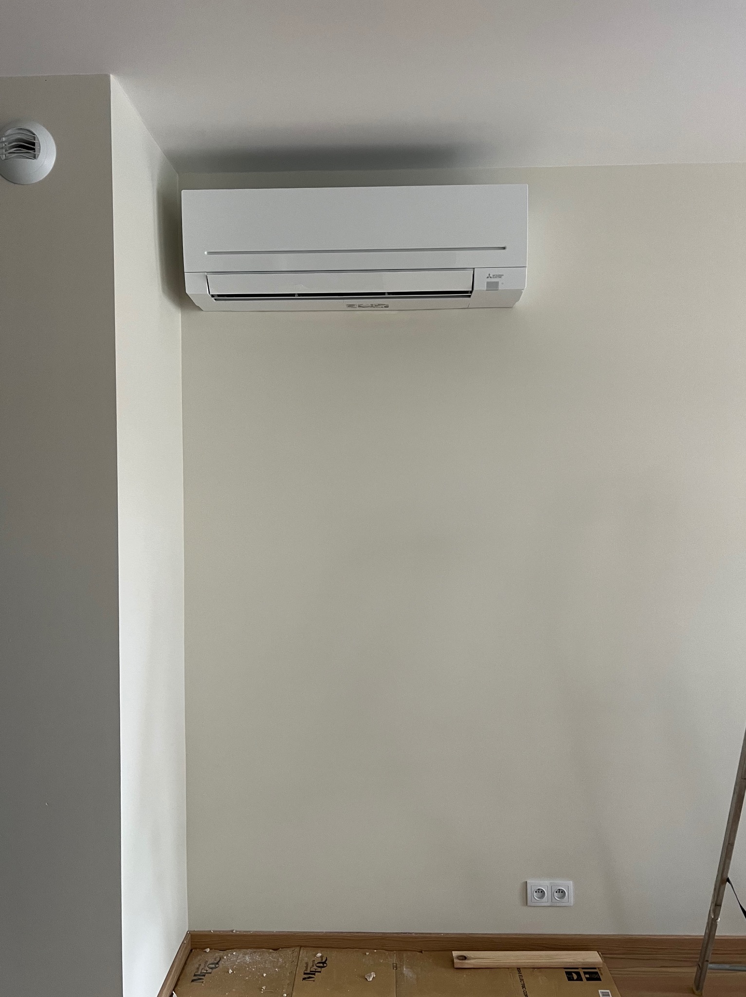 Biały klimatyzator Mitsubishi zamontowany na jasnej ścianie w pomieszczeniu. Widoczne gniazdka elektryczne i fragment drabiny. Minimalistyczny design.