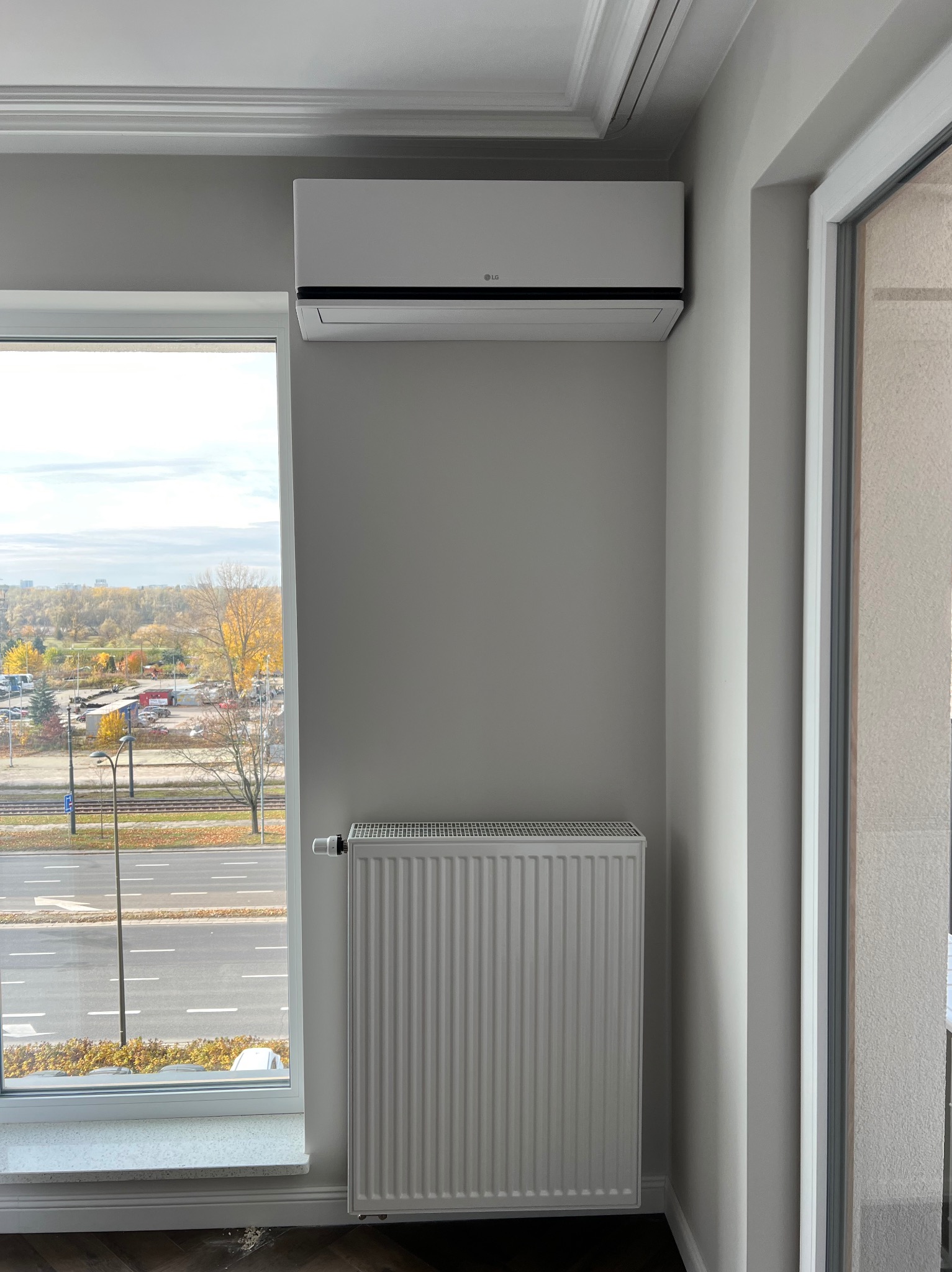 Biały klimatyzator LG zamontowany nad grzejnikiem w jasnym pokoju z widokiem na jesienny krajobraz miejski przez okno. Minimalistyczny design wnętrza.