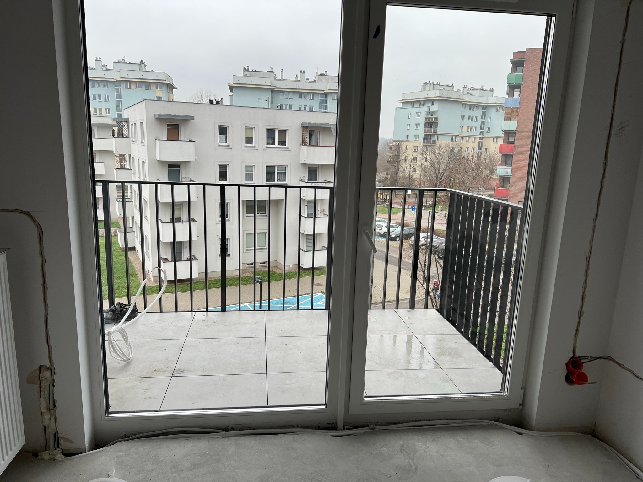 Widok z okna balkonowego na osiedle w Grodzisku Mazowieckim. Widoczne przewody elektryczne wystające ze ściany sugerują prace instalacyjne.