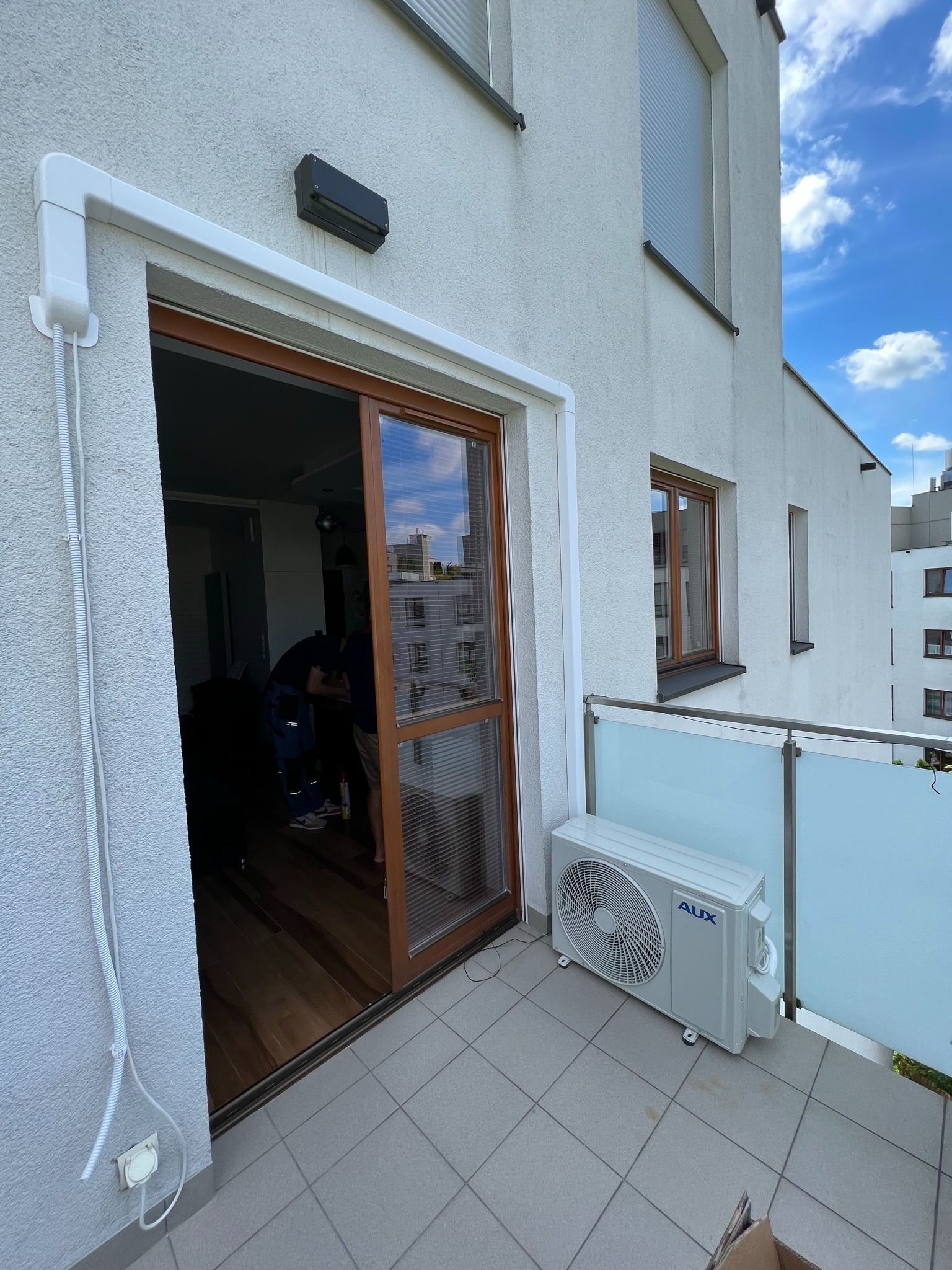 Jednostka zewnętrzna klimatyzatora marki AUX zamontowana na balkonie nowoczesnego apartamentowca, widoczne białe korytko instalacyjne na elewacji i pracownik wewnątrz mieszkania podczas instalacji.