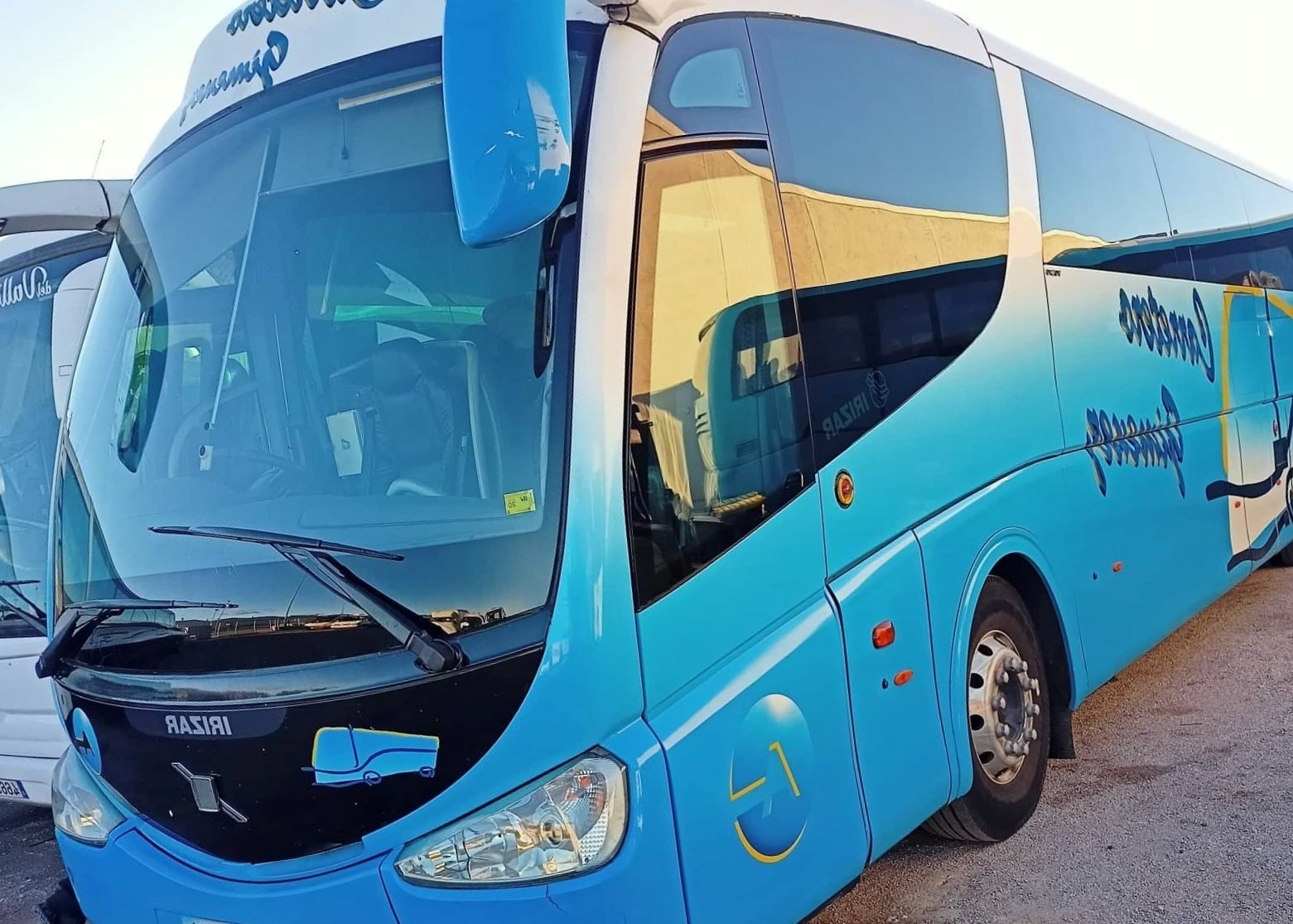 Niebieski autokar turystyczny z dużymi oknami i napisem firmowym, zaparkowany na placu, z widocznym odbiciem budynku w szybach.