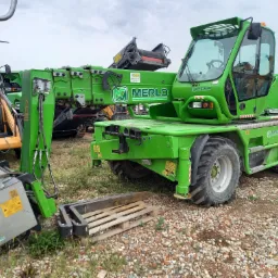 Zielona ładowarka teleskopowa Merlo 45.21 z podpiętą przystawką i paletą na żwirowym podłożu, widoczna w otoczeniu innych maszyn budowlanych.