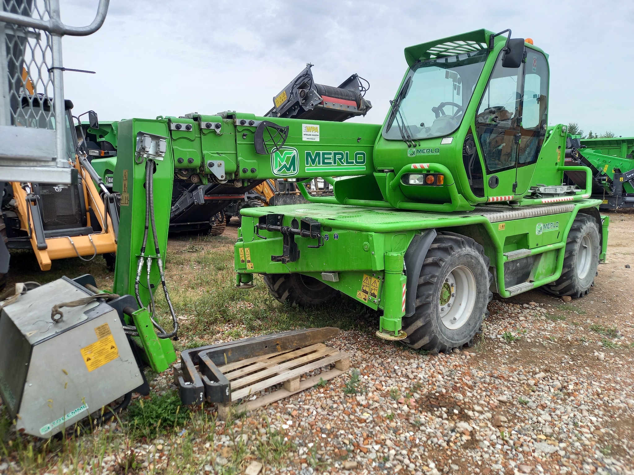 Zielona ładowarka teleskopowa Merlo 45.21 z podpiętą przystawką i paletą na żwirowym podłożu, widoczna w otoczeniu innych maszyn budowlanych.